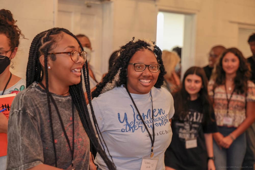 Two young ladies stand side by side, smiling together during an activity at a youth camp.